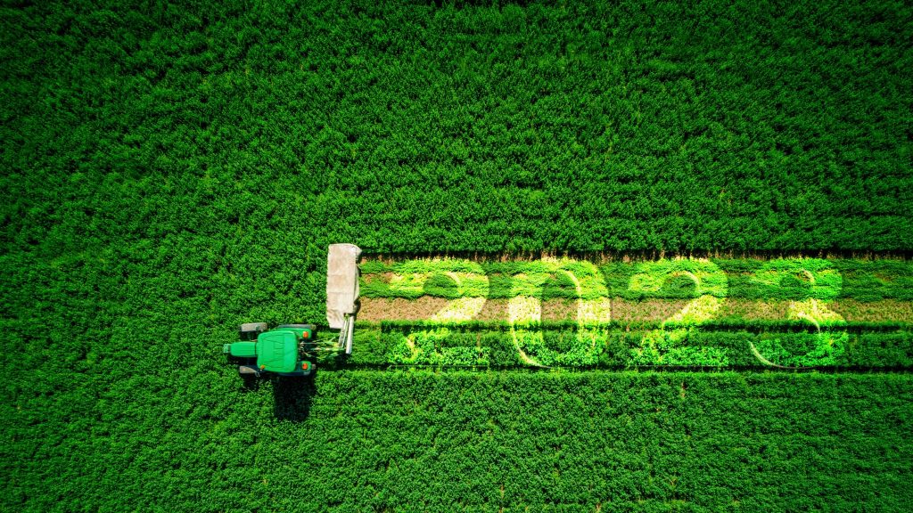 Une vue aérienne où un tracteur moissonne un champ vert et l'inscription "2023" apparait dans son sillage