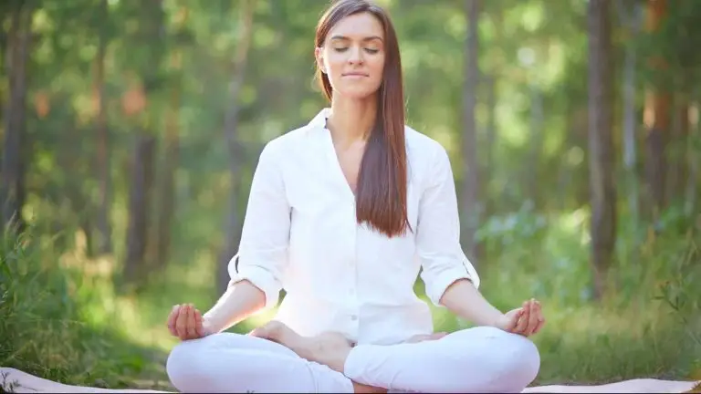 Une dame habillée en blanc est assise en tailleur sur un tapis de yoga