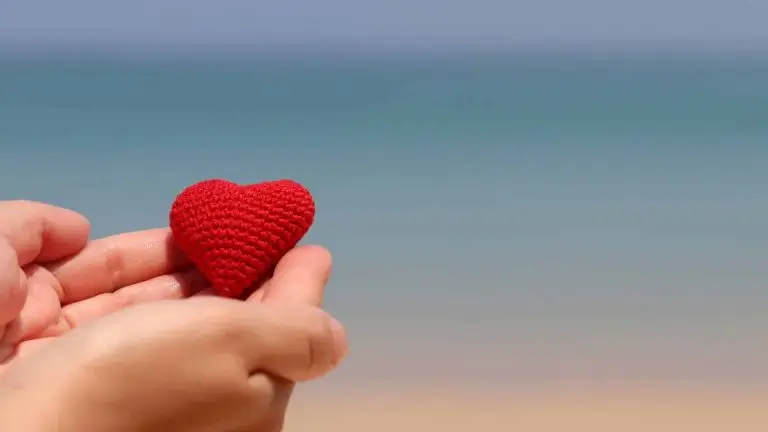 Une personne tient dans sa main un cœur rouge et derrière, il y a la plage et la mer