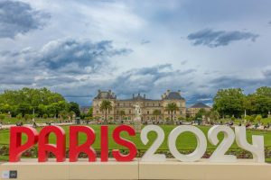 A promotional installation featuring the "PARIS2024" logo in bold red letters, set against a backdrop of a historic palace and gardens, under a cloudy sky.