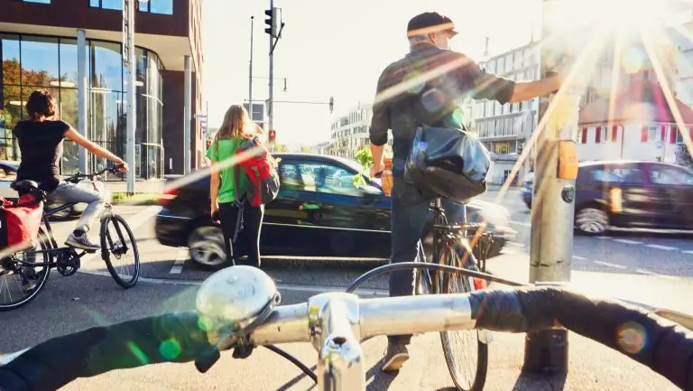 Il y a différents usagers de la route arrêtés à un feu rouge en ville : un piéton, des cyclistes et un automobiliste