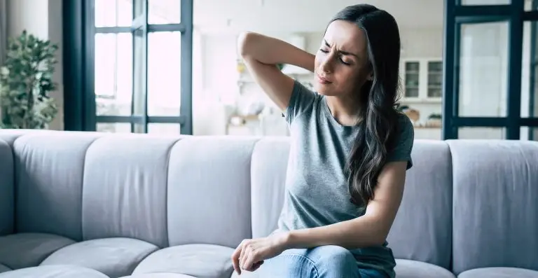 Une femme assise sur un canapé qui a des douleurs dans le dos se tient la nuque