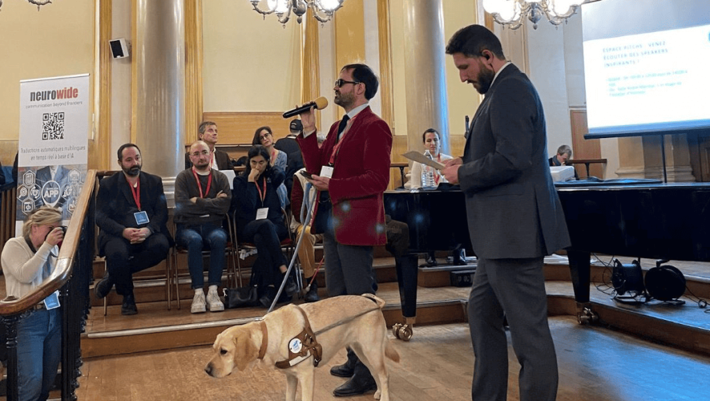 Thibaut de Martimprey chargé de projet du campus Louis Braille est en train de parler au micro. Son chien-guide se teint à ses côtés. Plusieurs personnes sont autour de lui.